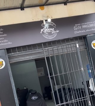 A frente da barbearia: letreiro escuro com o logo, o telefone e a lista de serviços sobre a porta de grade.