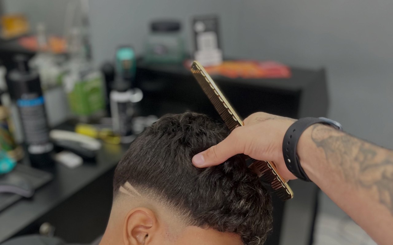 A mão do barbeiro com o pente levantando o cabelo no topo, a linha do degradê já feita na lateral.
